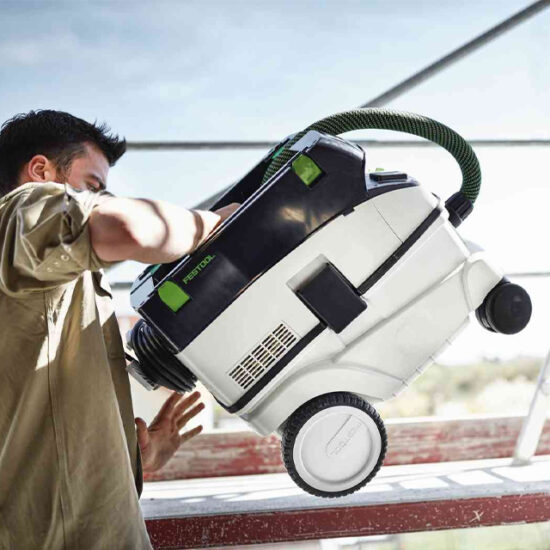 A person lifts a white and black Festool 574944 Mobile Dust Extractor CLEANTEC CTL 26 E AC 230V with large wheels and a green hose, standing outdoors near a metal structure.