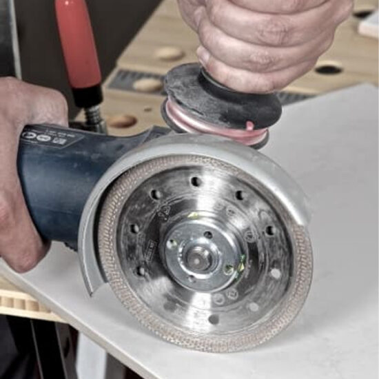 A person uses a Klingspor 331039 DT 900 FP Special 115mm diamond cutting blade with an angle grinder to cut white material, such as hard floor tiles or ceramics, on a workbench.