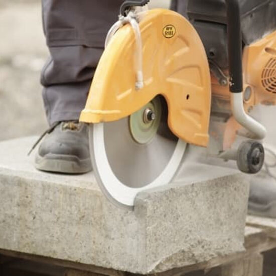 A person uses the Klingspor 336223 300 x DT 350 U Extra Large diamond cutting blades for construction materials in a yellow circular saw to cut a concrete slab, wearing protective work boots and pants as dust rises from the cutting edge.