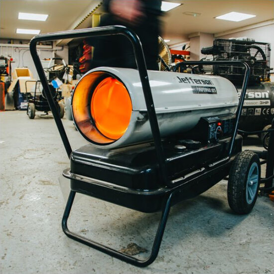 A Jefferson JEFHTRAD125TC Infrared 125 Heater with a glowing orange nozzle is positioned on a wheeled frame in a workshop, surrounded by tools and equipment.
