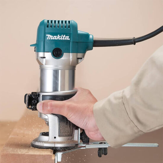 Using the Makita RT0702CX4/2 3/8 & 1/4 Router Trimmer, 710w 240v, someone shapes a wooden boards edge as wood shavings appear during trimming.