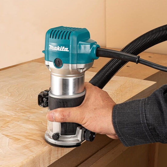 A person uses a Makita RT0702CX4/2 3/8 & 1/4 Router Trimmer 710w 240v to smooth the edge of a wooden board, attaching a vacuum hose for efficient dust collection.