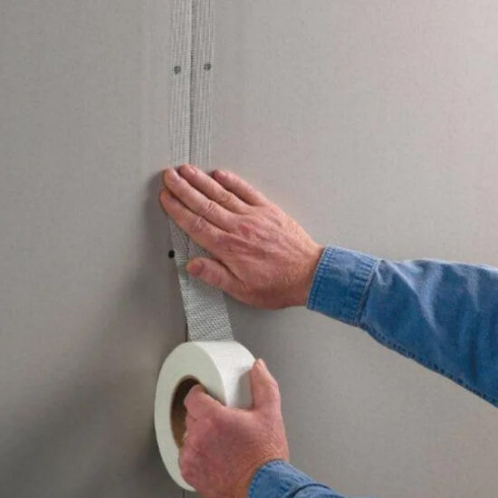 A person in a denim shirt applies a strip of mesh drywall tape vertically over the seam between two drywall panels on a gray wall.