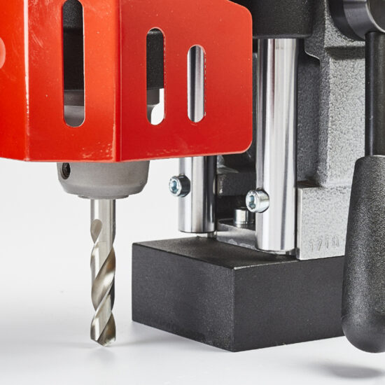 Close-up of a metal drill press with a spiral drill bit, featuring a red safety guard, metal rods, and a black base on a white background.