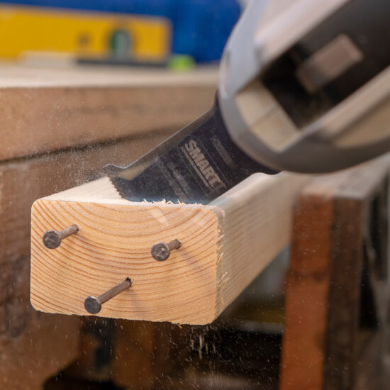 Using the Smart H3MAK Universal 3 Piece Blade Set, a reciprocating saw cleanly cuts a wooden beam with three nails, producing wood dust and showcasing the blades precision and effectiveness on tough materials.