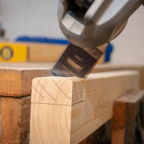 A close-up shows a power saw with the Smart H3MAK Universal 3 Piece Blade Set cutting wood, sending sawdust flying. Measurement markings cover the wood, and a level tool is visible in the blurred background.