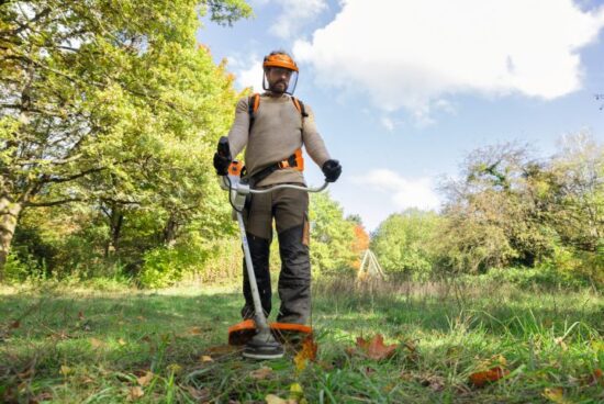 Wearing Stihl 0088458010 FS3PROTECT Protective Trousers, a person uses a brushcutter to trim grass in an outdoor area surrounded by trees under a partly cloudy sky.