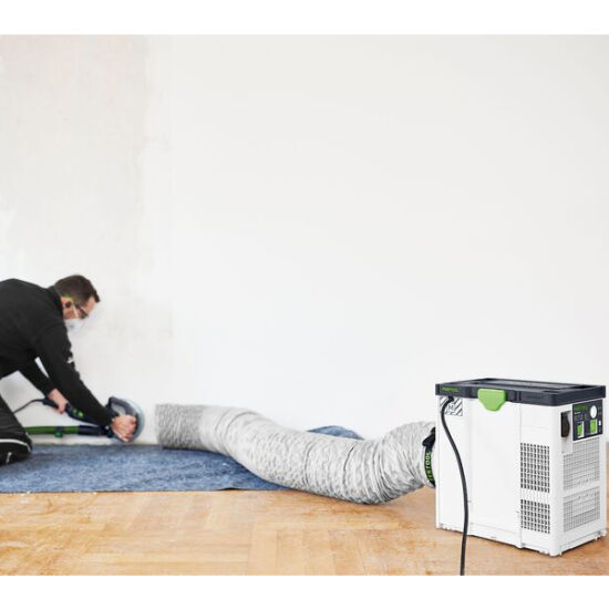 A person wearing safety goggles uses a tool on a blue carpet, connected by a large hose to a Festool Air Purifier SYS-AIR M placed on the wooden floor.
