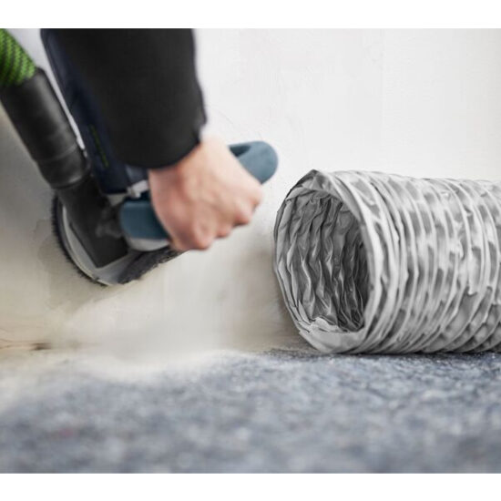 A person uses a power tool to cut a hole in a white wall near a flexible silver air duct on blue carpet, while the Festool Air Purifier SYS-AIR M helps keep the air clean during the process.