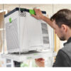 A man mounts the Festool Air Purifier SYS-AIR M, which is white, onto a ceiling platform in a workshop, securing it by its green handle with one hand.