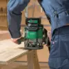 Wearing blue work clothes, a person uses a Hikoki HIKM3612DAJP M3612DA/JPZ 1/4 & 1/2in Multi-Volt Router 36V with a 2.5Ah Li-ion battery on the edge of a wooden table in a workshop.