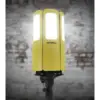 A yellow Firthfull FPPCENTAURL Centaur Heavy-Duty 200W LED Site Light (110V) with three illuminated panels stands on a black frame, set against a blurred brick wall background.