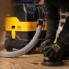 A person wearing gloves uses a handheld electric sander attached to a yellow and black dust extractor, sanding a wooden surface indoors.