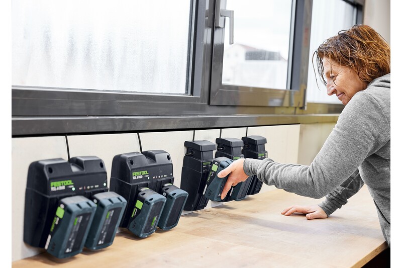 A person places a battery into one of several Festool battery chargers mounted on a wooden workbench below a window. The chargers are lined up and plugged in, with multiple batteries already charging.