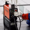 In a busy workshop, a person wearing protective gloves and a Lorch-branded sleeve adjusts the controls of a red and black Lorch MicorTIG 200 BasicPlus welding machine, which shows 81A on its digital display.