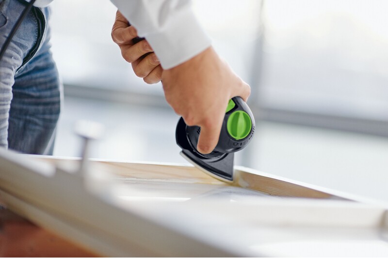 A person uses a small electric sander with a green accent to smooth the edge of a wooden surface, focusing on the sanding process.