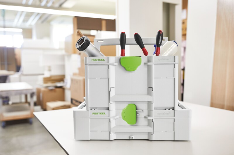 A white and green Festool toolbox with compartments and various tools, including screwdrivers and tape, is placed on a table in a bright, modern workspace with shelves and boxes in the background.