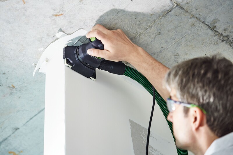 A person wearing safety glasses uses a power sander with a green hose attachment to smooth a white, curved surface under a concrete ceiling.
