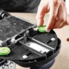 A close-up of a person’s hand adjusting a green knob on a metal and black power tool placed on a wooden surface with holes.