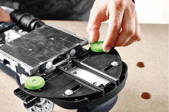 A close-up of a person’s hand adjusting a green knob on a metal and black power tool placed on a wooden surface with holes.