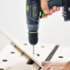 A person uses a cordless Festool power drill to drill a hole into a piece of wood placed on a workbench. The focus is on the drill and the individuals hand.