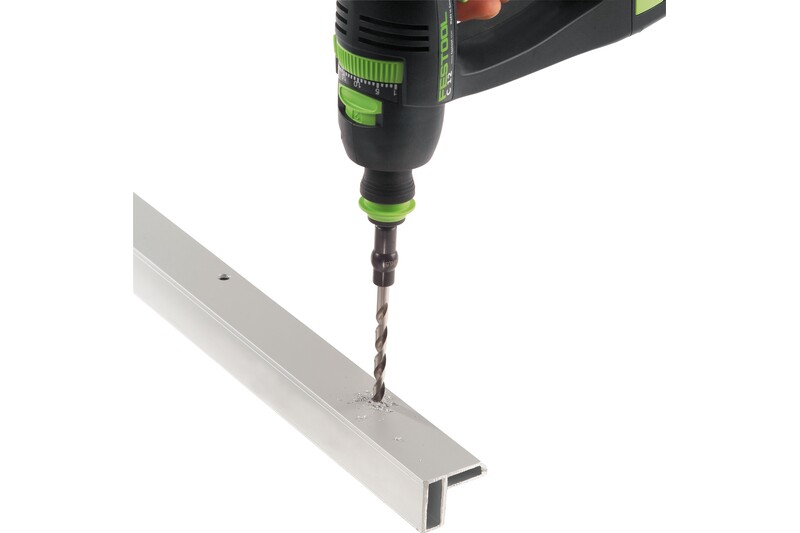 A close-up of a power drill with a green and black handle drilling a hole into a piece of light-colored metal, with aluminum shavings scattered around the drill bit.