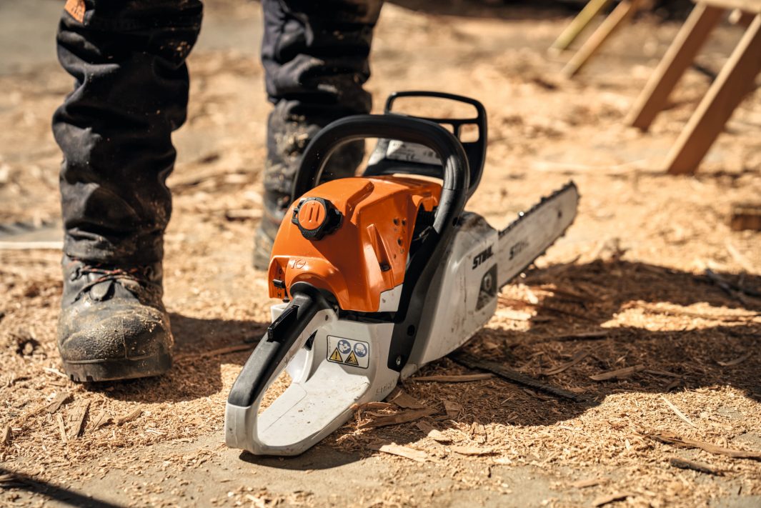 A person in work boots stands by an orange and white Stihl chainsaw with wood shavings outdoors, focusing on tool management.