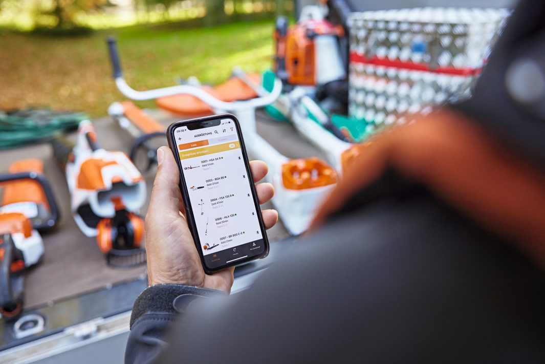 A person stands by Stihl outdoor power tools, holding a smartphone with a list; tools rest on a table before a blurred green background.