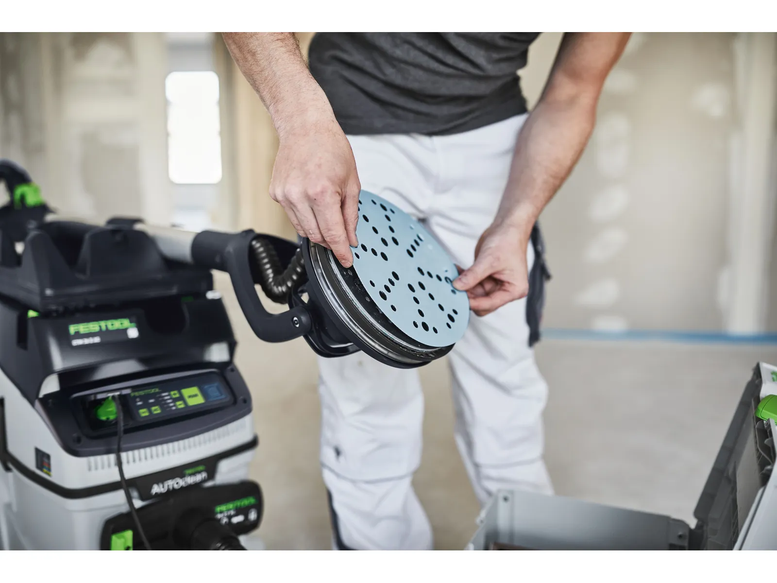 A person in work clothes attaches a sanding disc to an electric sander in a room under construction, with tools visible nearby.