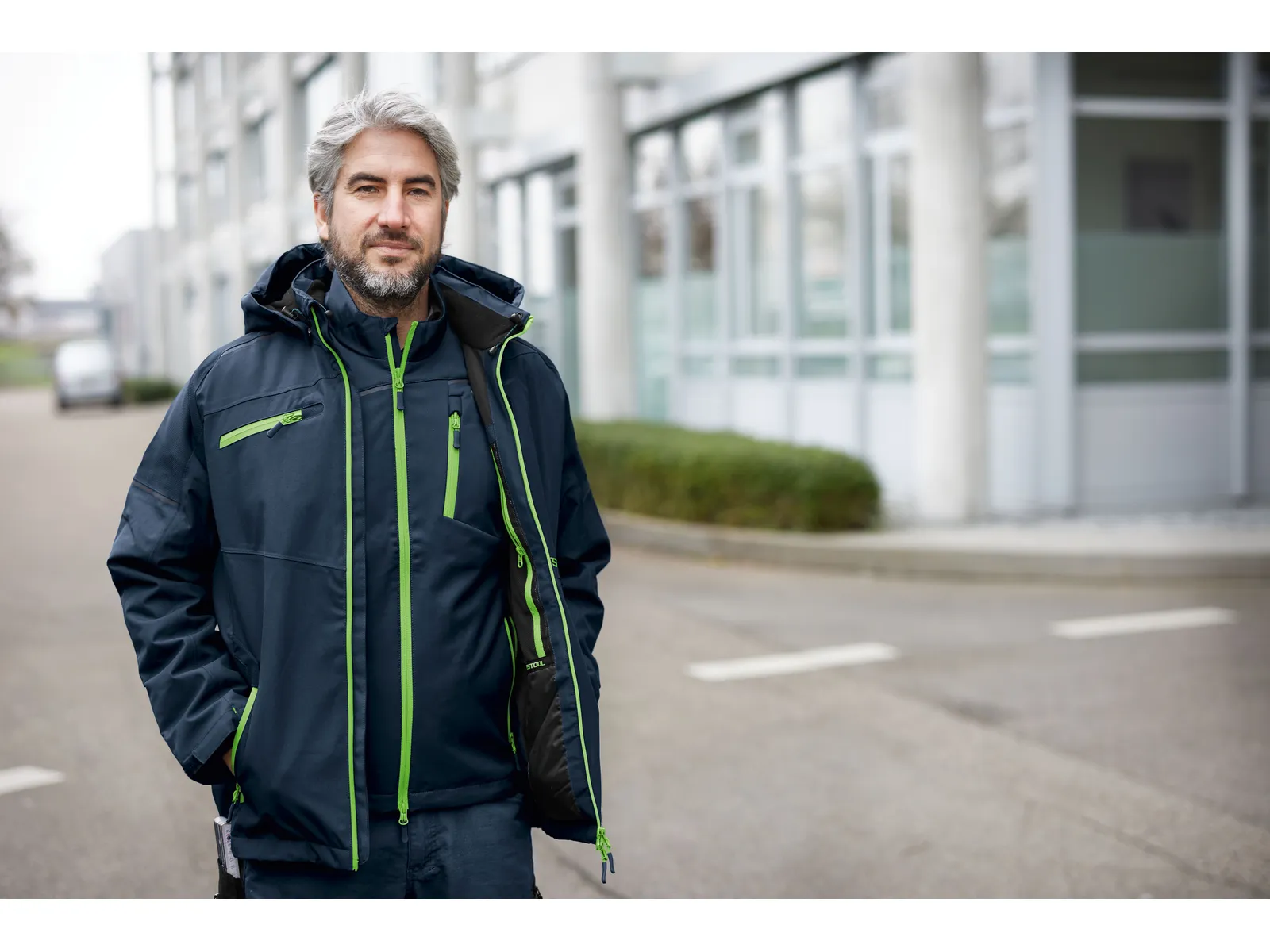 A man with gray hair and a beard stands outside in a dark blue jacket with green zippers, in front of a modern building.