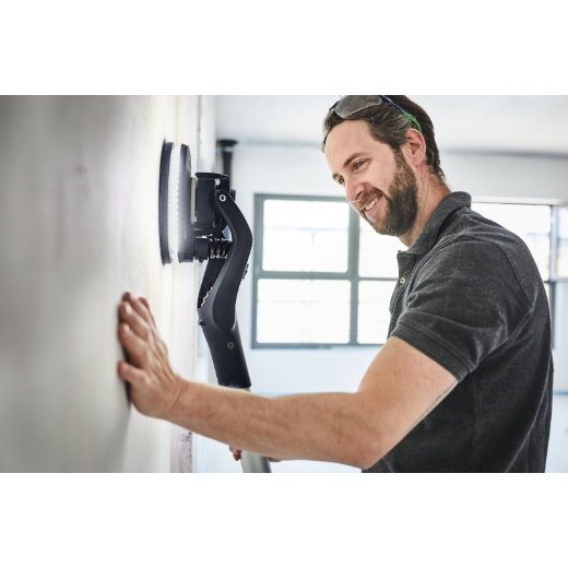 A man in a dark polo shirt and safety glasses smiles while sanding a white wall in a well-lit room with a long-reach sander.