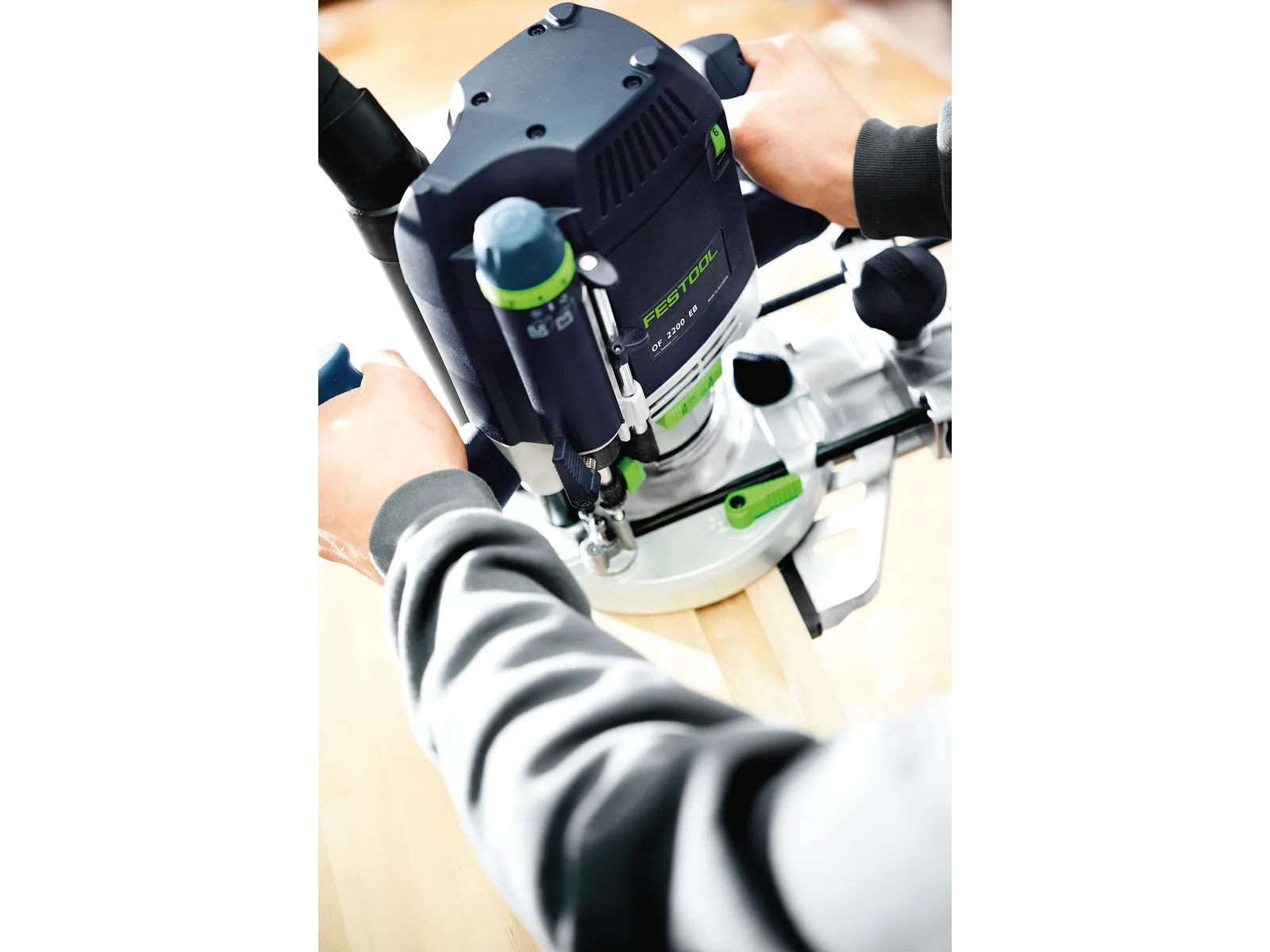 Person using a Festool power router to shape the edge of a light wooden board, hands steadying both the tool and the board.