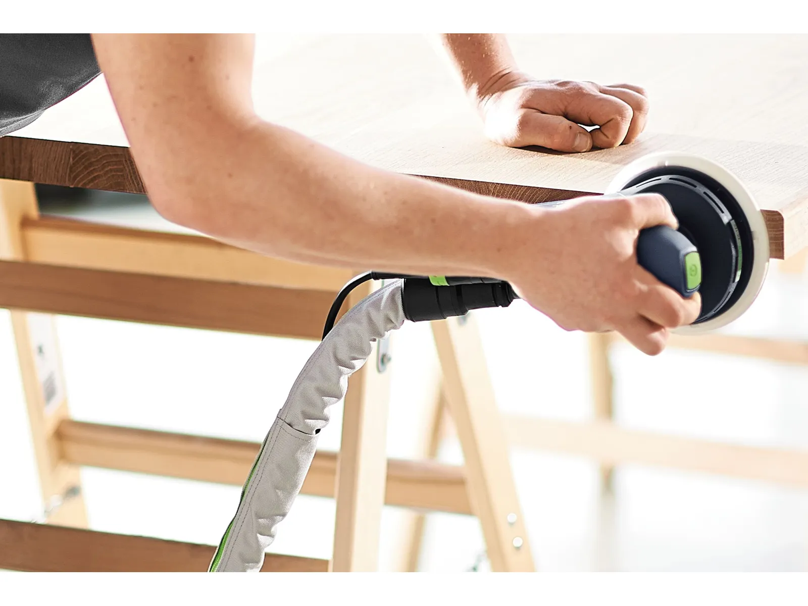 A person uses an electric sander to smooth a wooden plank on a sawhorse, holding the sander in one hand and steadying the wood.