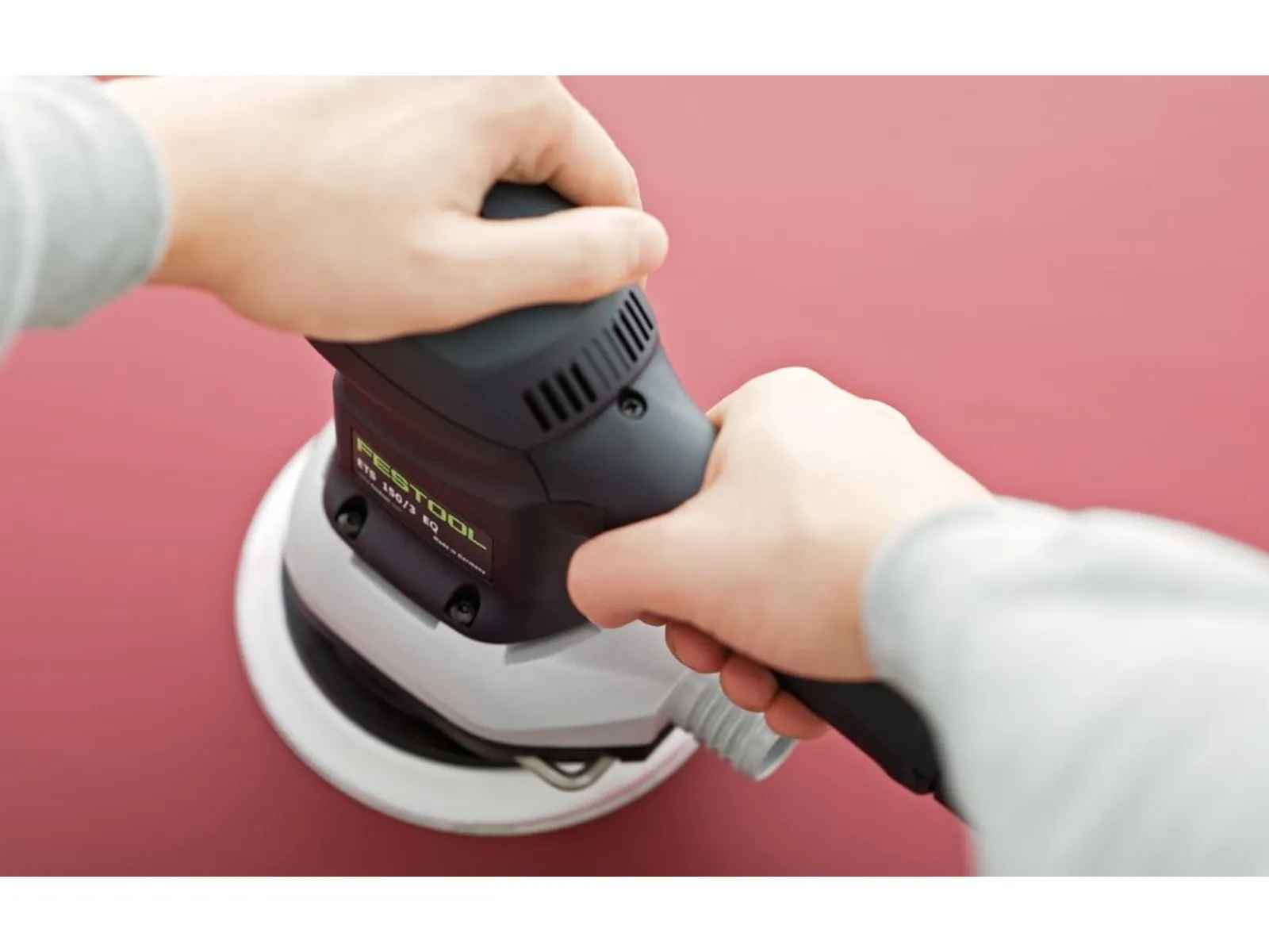 Close-up of two hands holding and operating a black and gray electric sander on a flat red surface.
