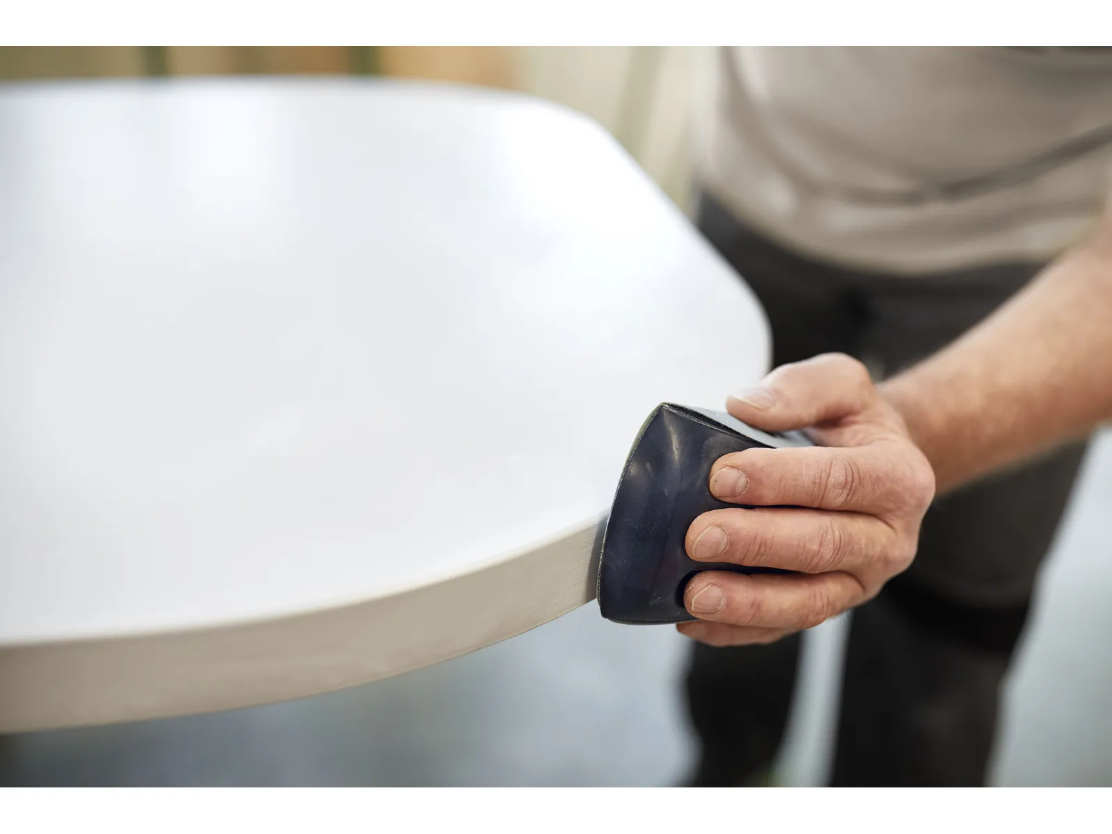 A person sands the edge of a white tabletop with a sanding block, paying close attention to smoothing and finishing the surface.
