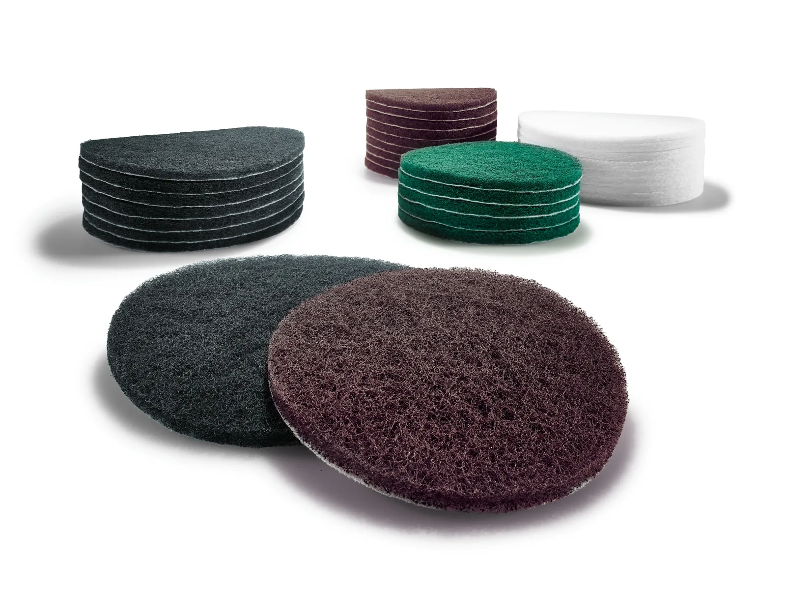 Stacks of round scrubbing pads in black, brown, green, and white on a white surface; two pads lie flat in the foreground.
