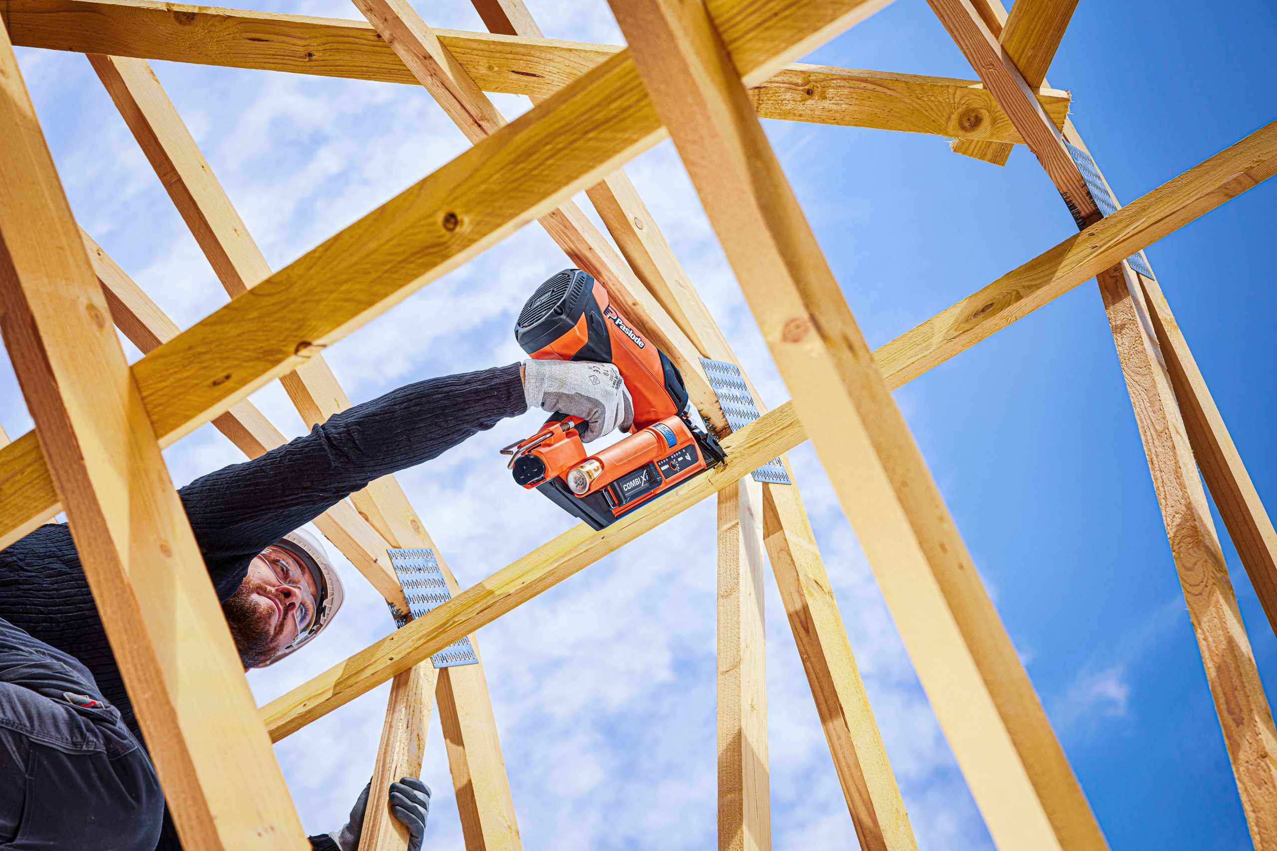 A construction worker secures roof frame beams with a Paslode Combi Xi tool under a blue sky, viewed from below, wearing gloves.