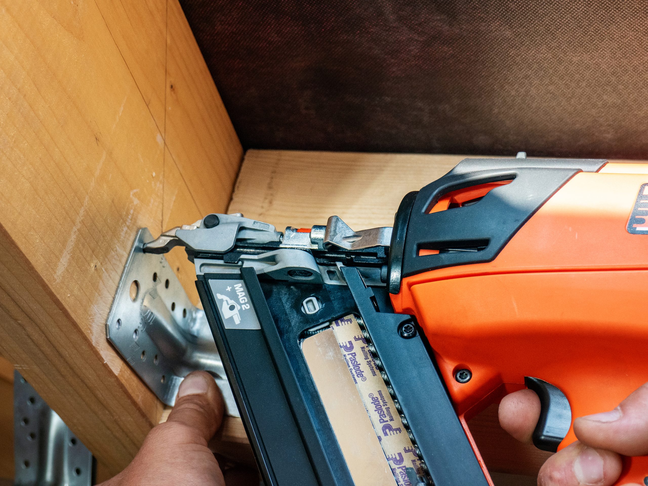 A close-up of a person using an orange Paslode Combi Xi One Tool to fasten a metal bracket to a wooden frame, holding it steady.