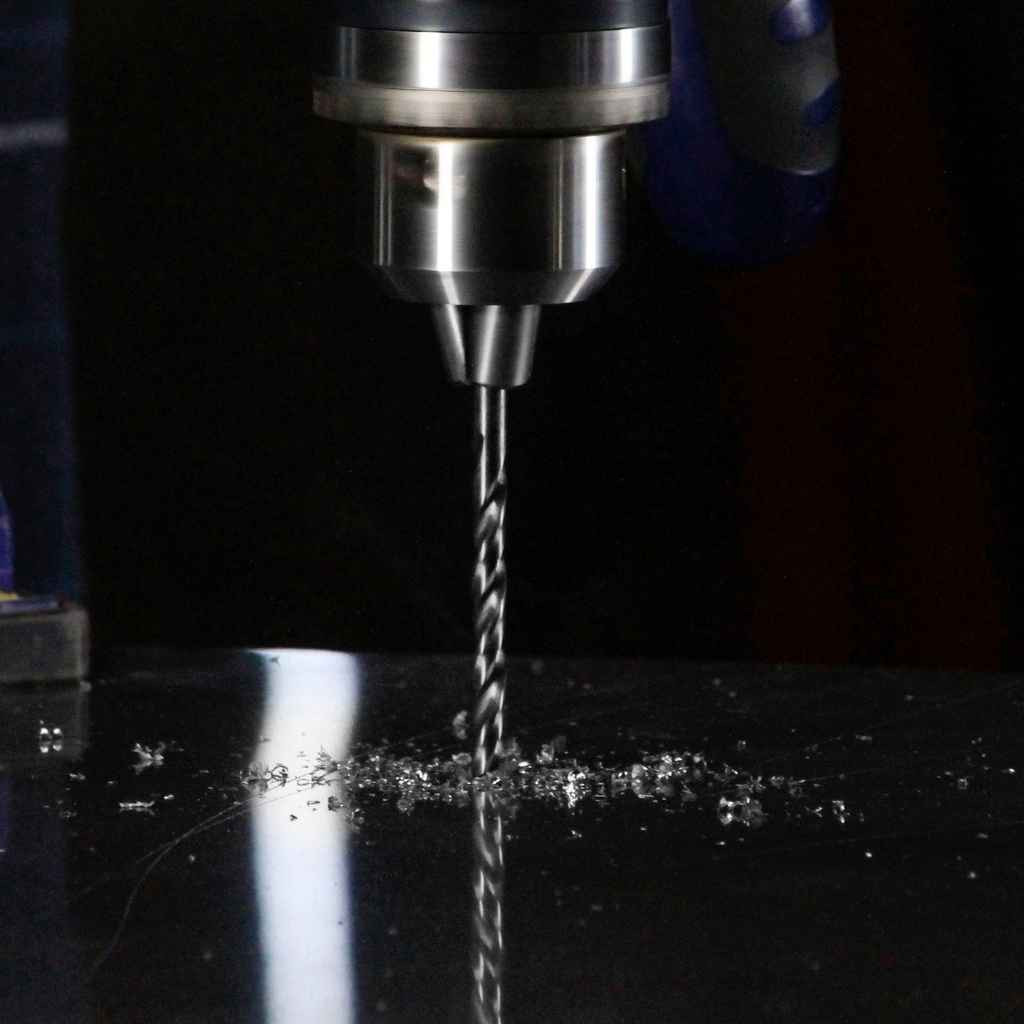A close-up of a 1.5mm Hss metric drill bit mounted on a drill press making a hole in metal, with shavings around the area.