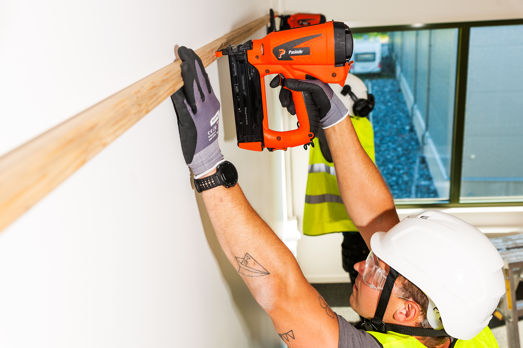 A construction worker in safety gear uses a nailer to secure wood to a white wall indoors; another worker is in the background.