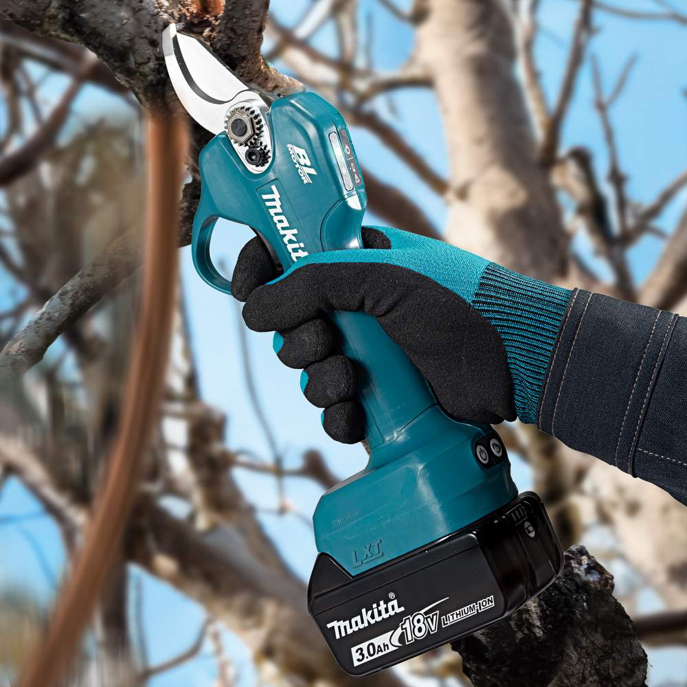 A gloved hand uses a Makita DHP487SF 18v Combi Drill with 3ah battery to prune a tree branch on a clear day with bare branches.