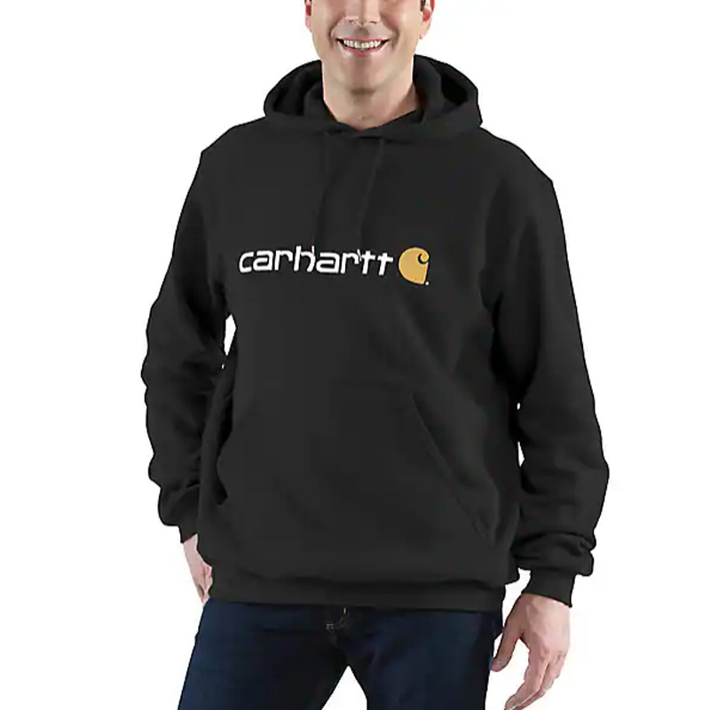 A person in a black Carhartt TS0074 Loose Fit Midweight Sweatshirt with a white logo and yellow emblem smiles, hands in pocket.