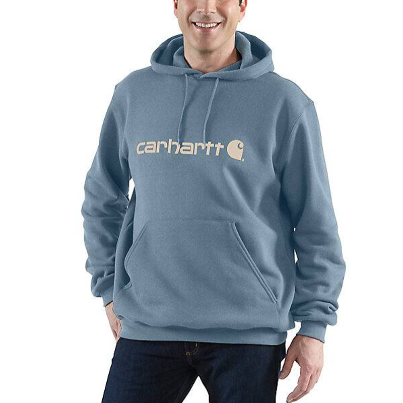 A smiling person wears a green Carhartt loose fit sweatshirt with a front pocket and beige logo on the chest, looking comfortable.