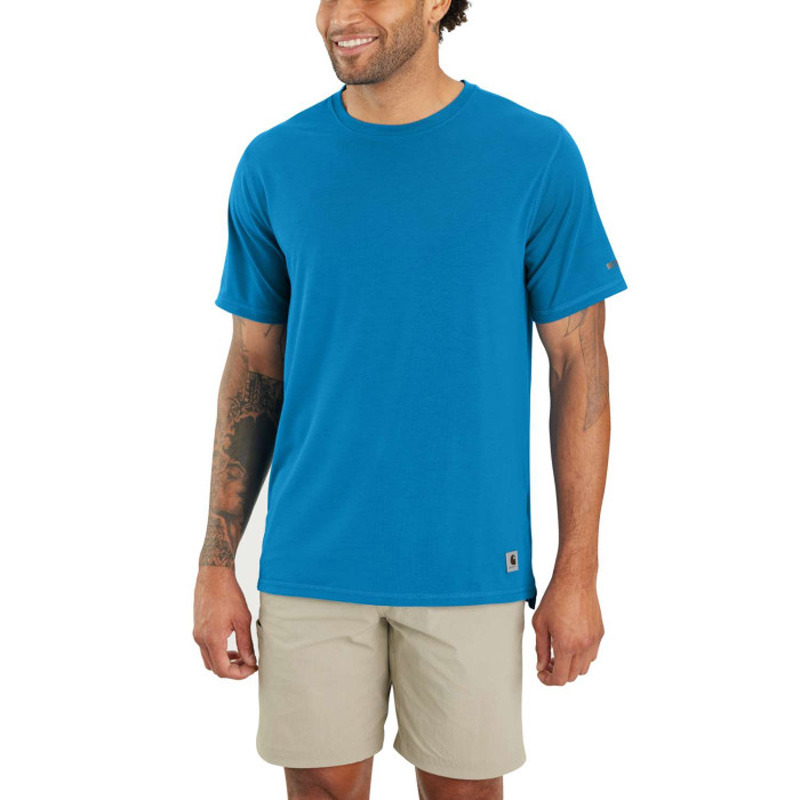 A man stands smiling with his arms relaxed at his sides, wearing a marine blue Carhartt t-shirt and light beige shorts.
