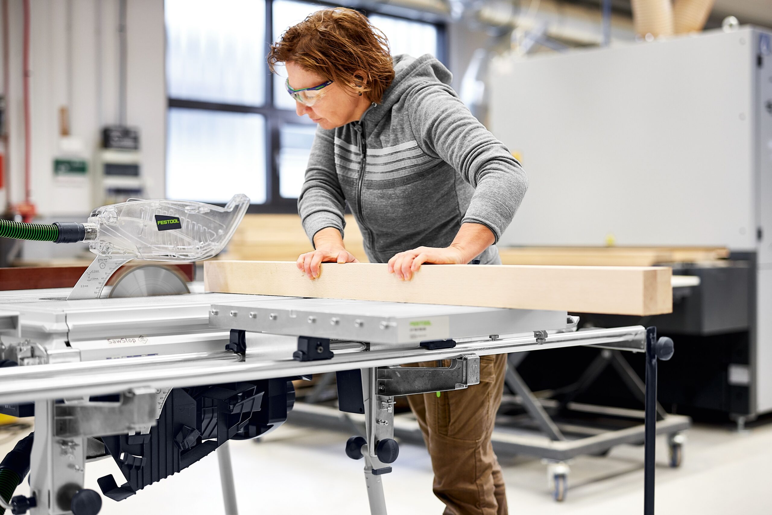 In a bright, modern workshop, a person in safety glasses and a gray hoodie uses the Festool Sliding Table to cut a wooden board.