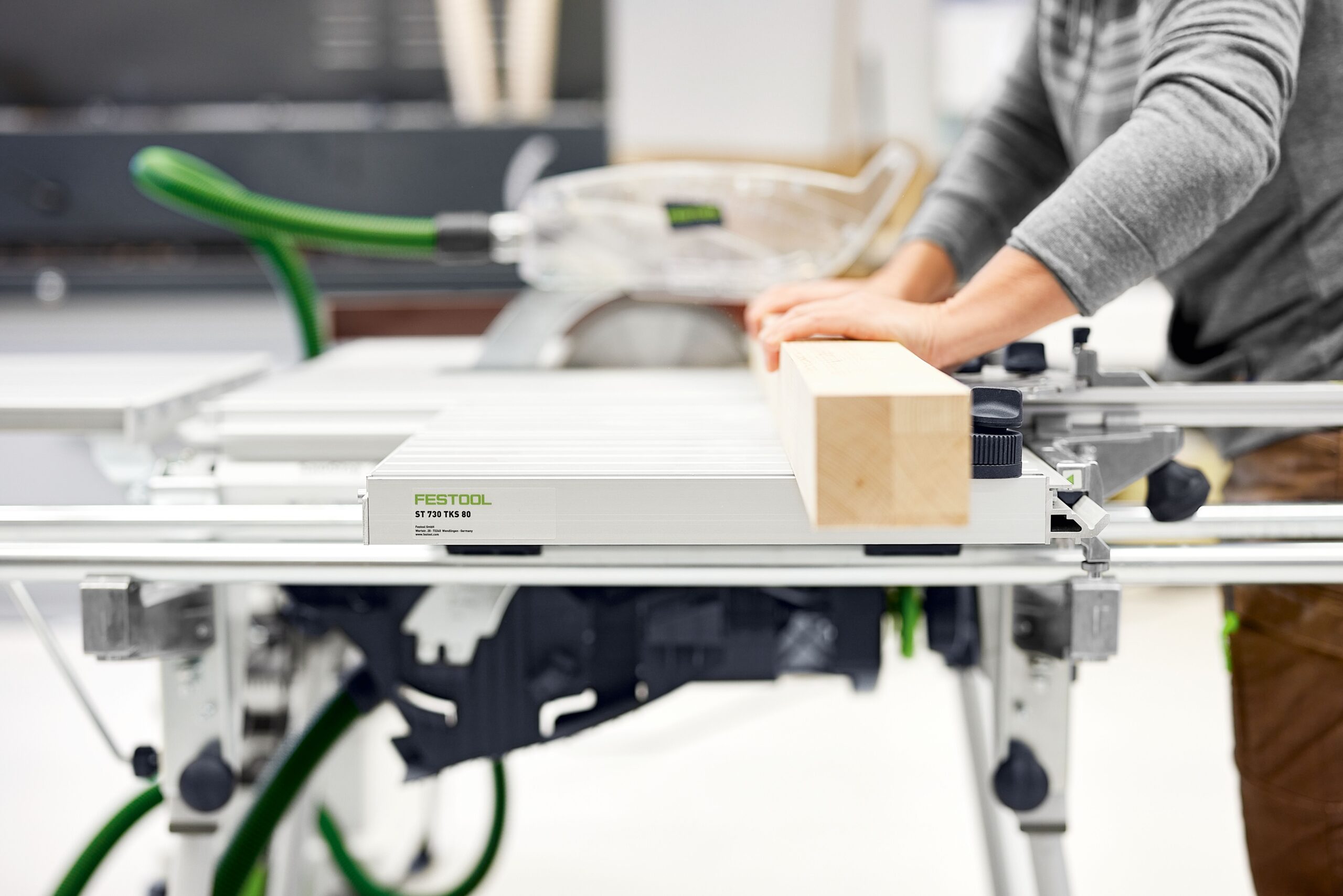 In a workshop, a person in a gray shirt uses a Festool table saw with sliding table to guide a wooden plank, blade visible behind.