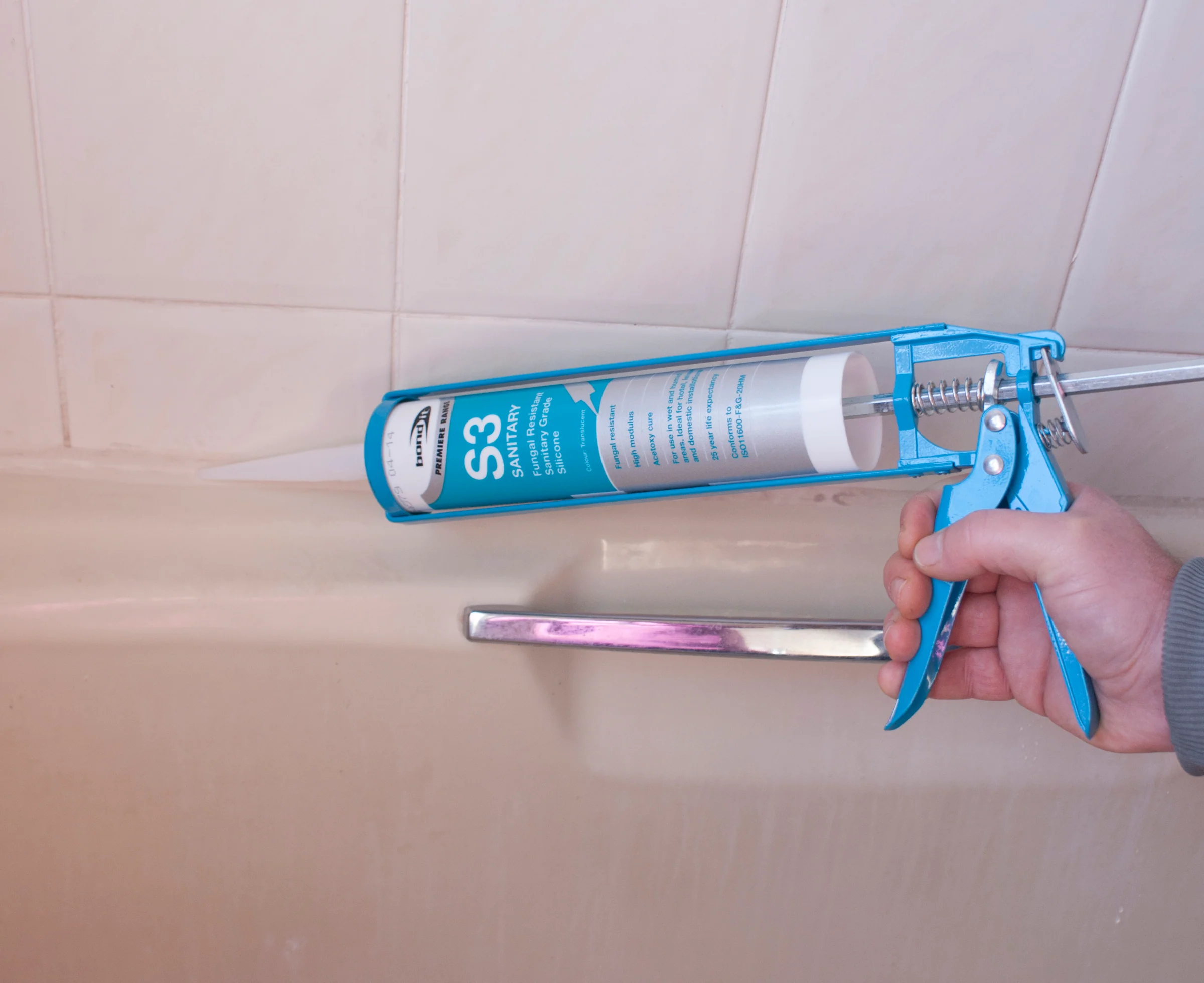 A person uses a caulking gun to apply white silicone sealant along the edge of a bathtub where it meets the tiled wall.