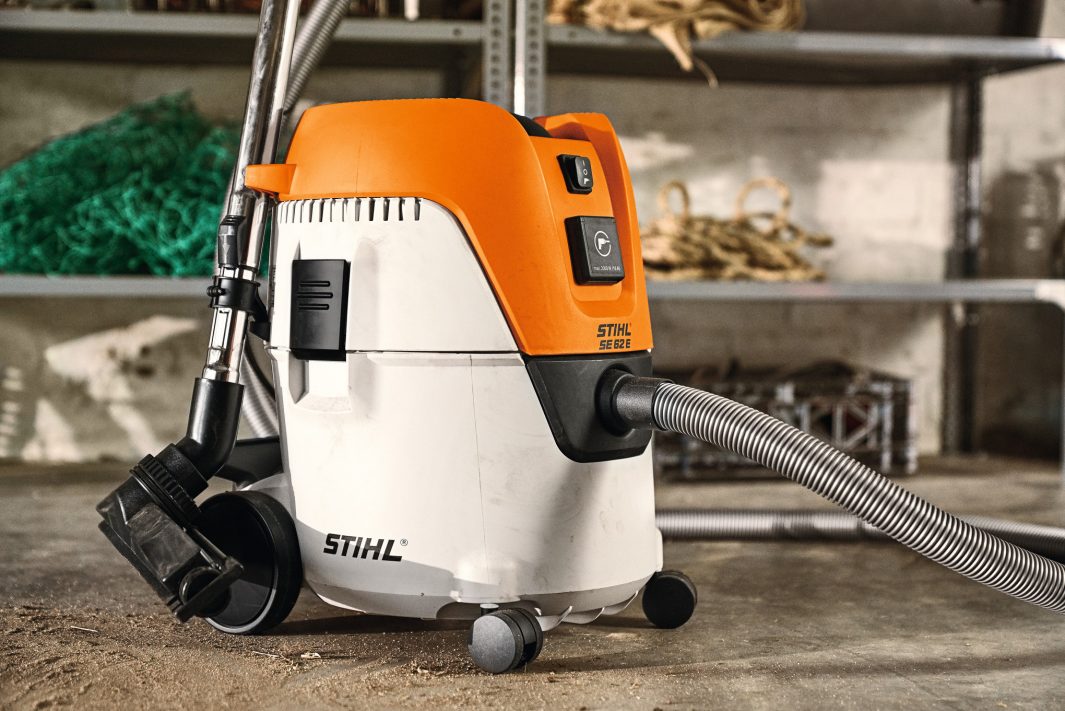 A Stihl SE 62 Vacuum Cleaner with an orange and white body and a silver hose stands on a workshop floor, shelves and tools behind.