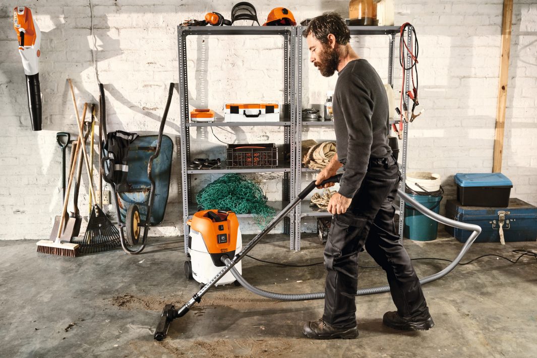 A man in work clothes cleans a concrete workshop floor with a Stihl SE 62 Vacuum Cleaner. Shelves with tools and supplies are behind him.