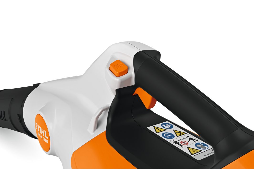 Close-up of a STIHL leaf blower handle with an orange power button, safety labels, and black grip on a white and orange body.